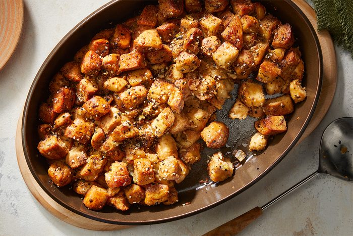 A brown oval baking dish filled with golden-brown, seasoned bread cubes, resembling baked stuffing or croutons, sits on a light countertop next to a serving spoon.