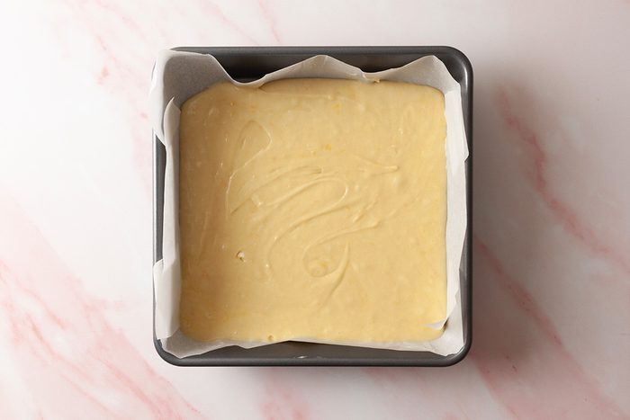A square baking pan lined with parchment paper, filled with smooth, light yellow cake batter, placed on a light pink and white marbled surface.