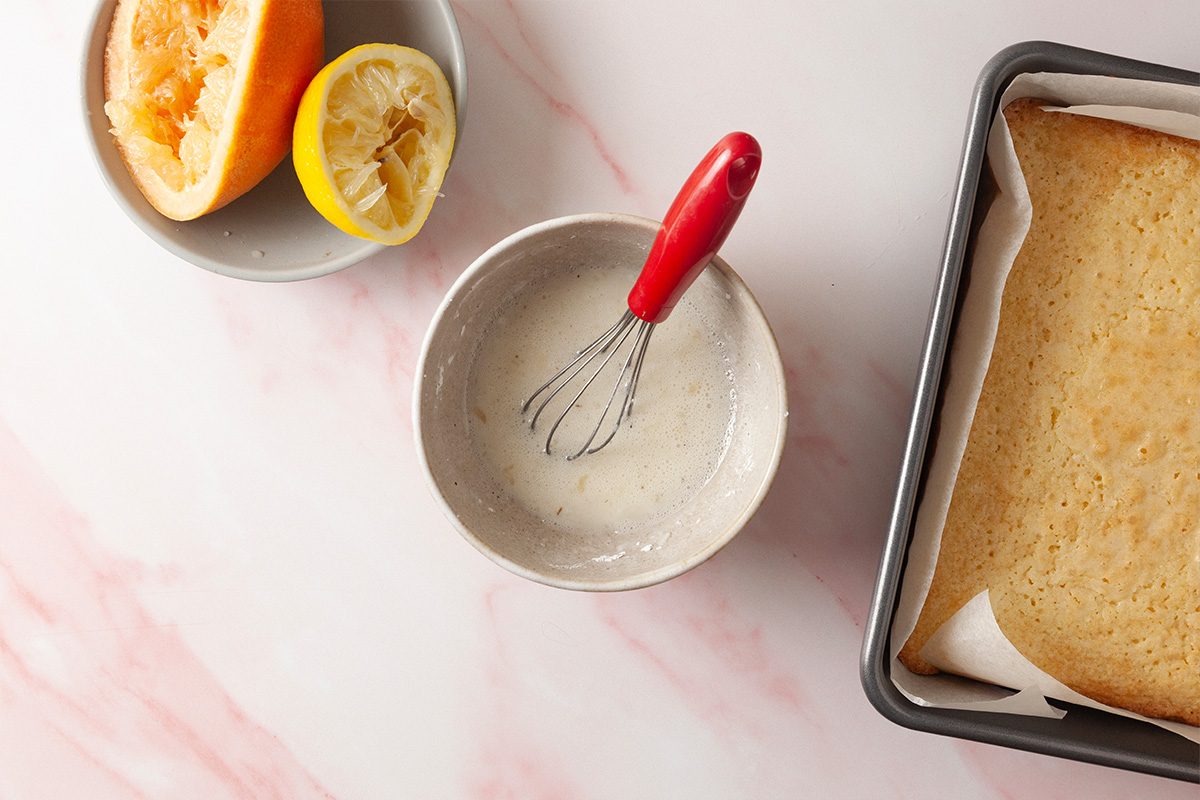A bowl with glaze and a whisk, a plate with squeezed grapefruit and lemon halves, and a baking pan with a sheet cake on a light pink marble surface.