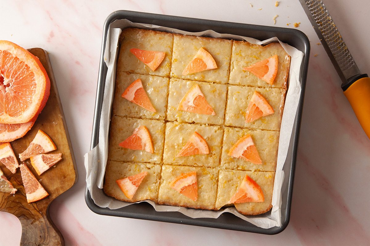 A square baking pan lined with parchment paper holds twelve grapefruit dessert bars, each topped with a small piece of fresh grapefruit. A zester and sliced grapefruit are nearby on a wooden board.