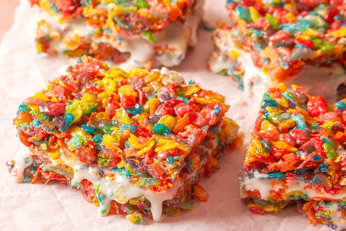 Close-up shot of Fruity Pebbles Milk 'n' Cereal Bars on parchment paper; showing colorful cereal squares with creamy white filling oozing between the layers