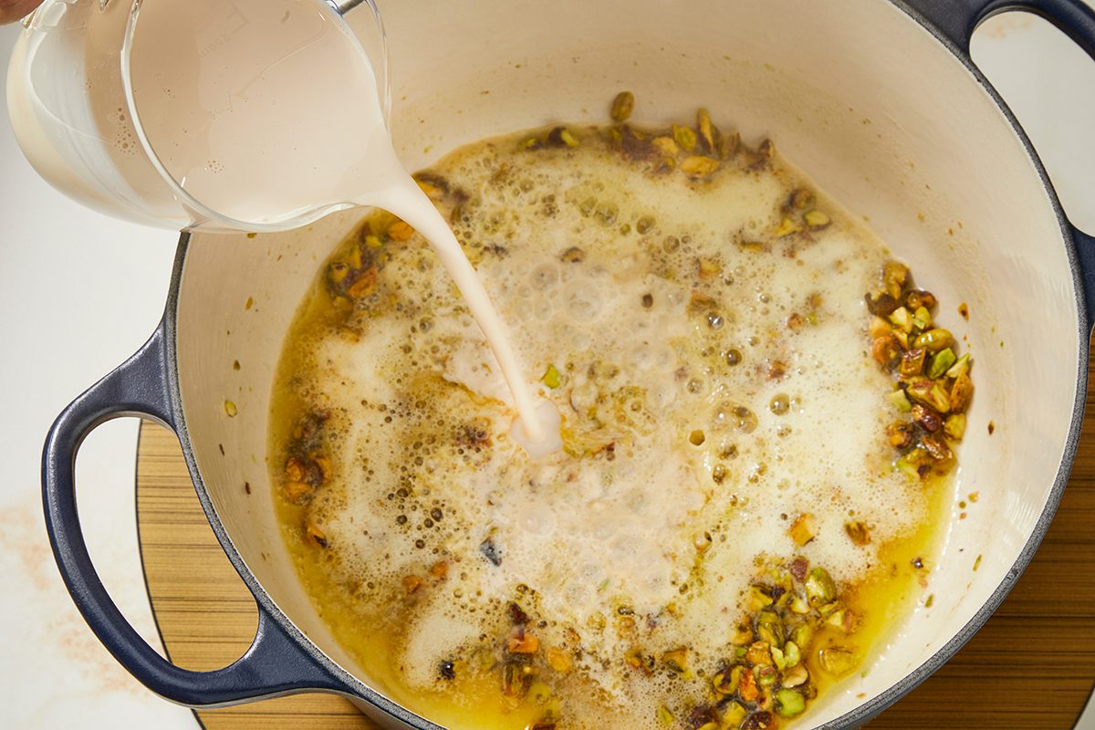 A hand pours a creamy liquid from a pitcher into a blue pot containing bubbling butter and chopped nuts on a stove.