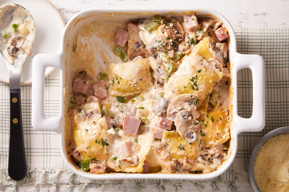 A white baking dish filled with creamy baked ravioli, visible pieces of ham, mushrooms, and green herbs on top, rests on a checkered cloth next to a serving spoon.