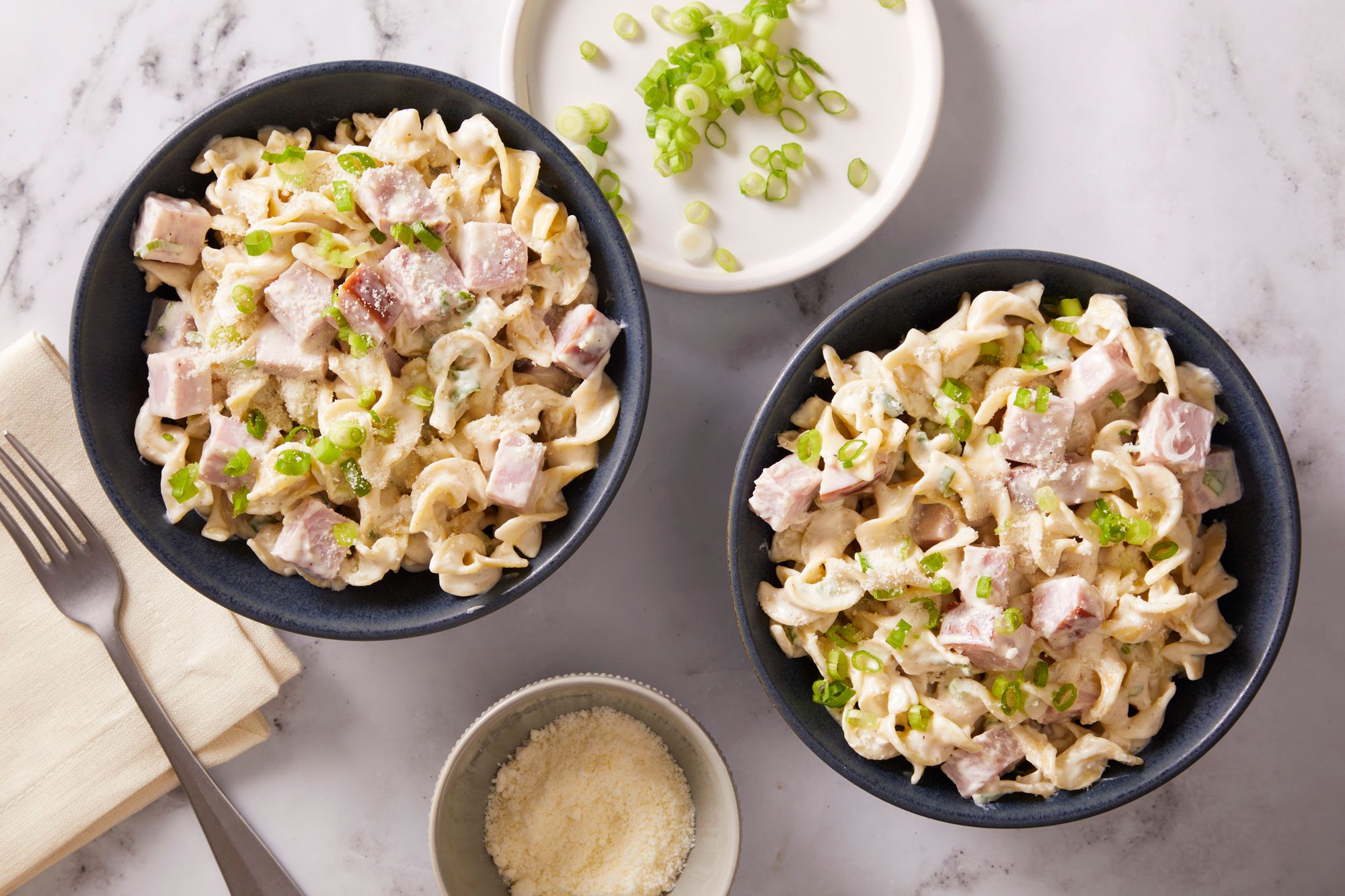 Overhead shot of Two small black bowls of Hurry-Up Ham N Noodles, accompanied by a small bowl of cheese and a fork.
