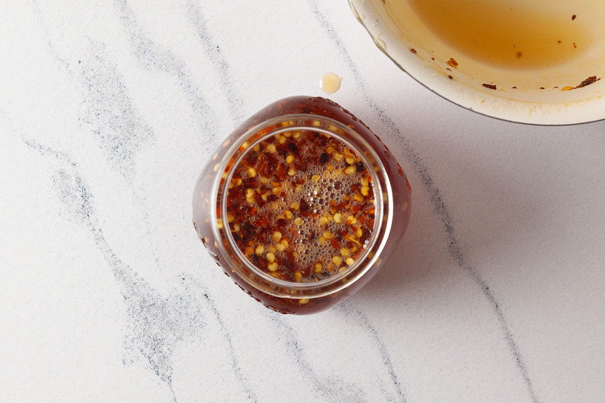 A small glass jar filled with a mixture of chili flakes and oil sits on a white marble surface next to a partially visible bowl containing a light brown liquid.
