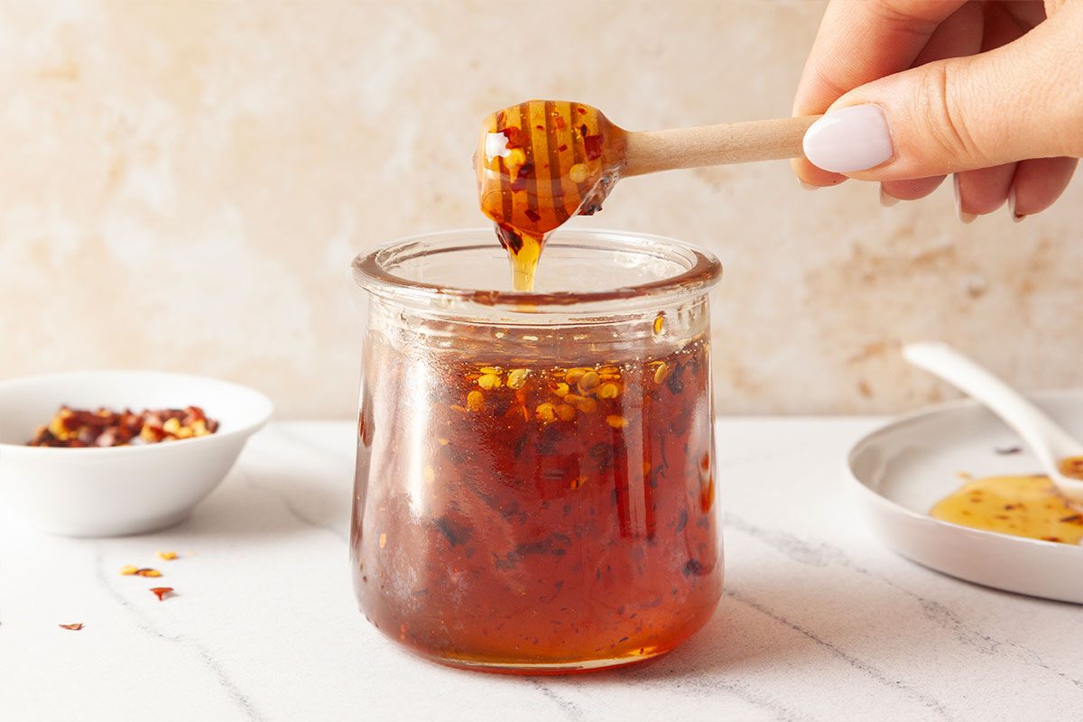 A hand holds a honey dipper over a glass jar filled with chili-infused honey. Two small white dishes, one with chili flakes and another with honey, sit nearby on a white surface.