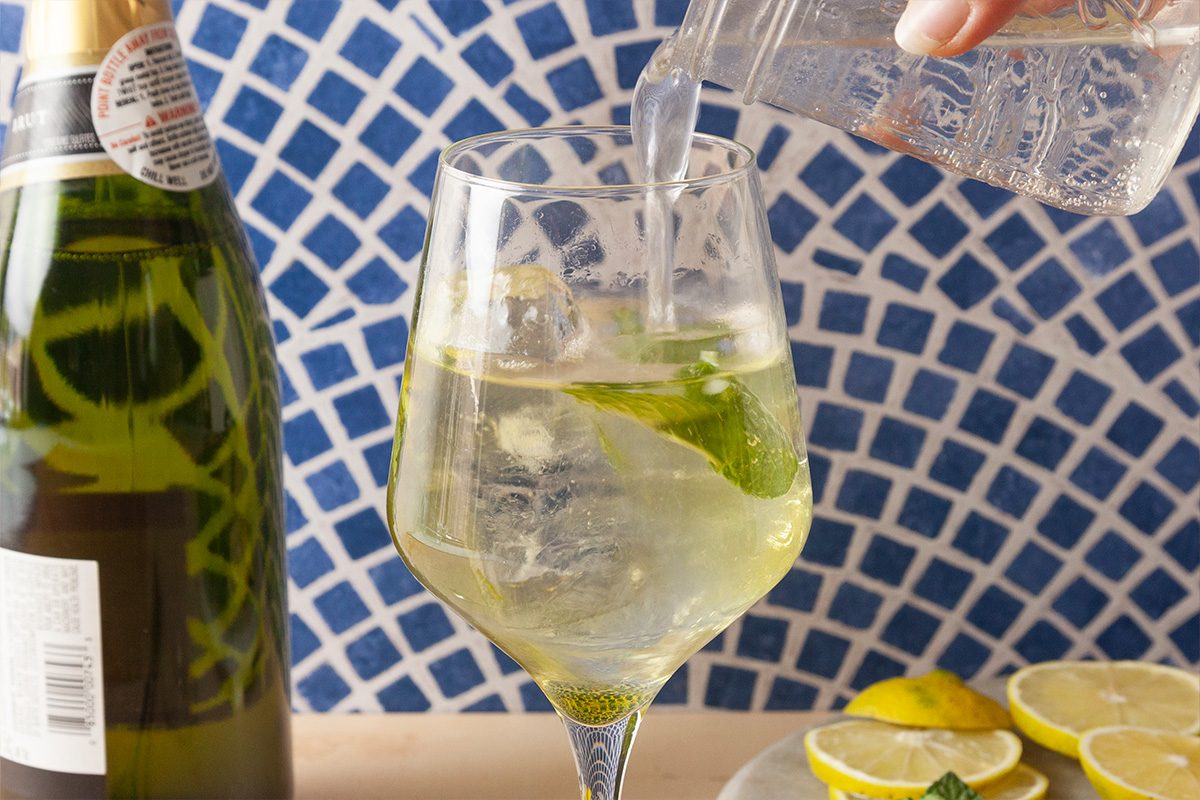 A glass of sparkling beverage with ice and mint is being poured into a wine glass. A bottle is next to the glass, and sliced lemons are on a plate in the background, with a blue and white tiled wall behind.