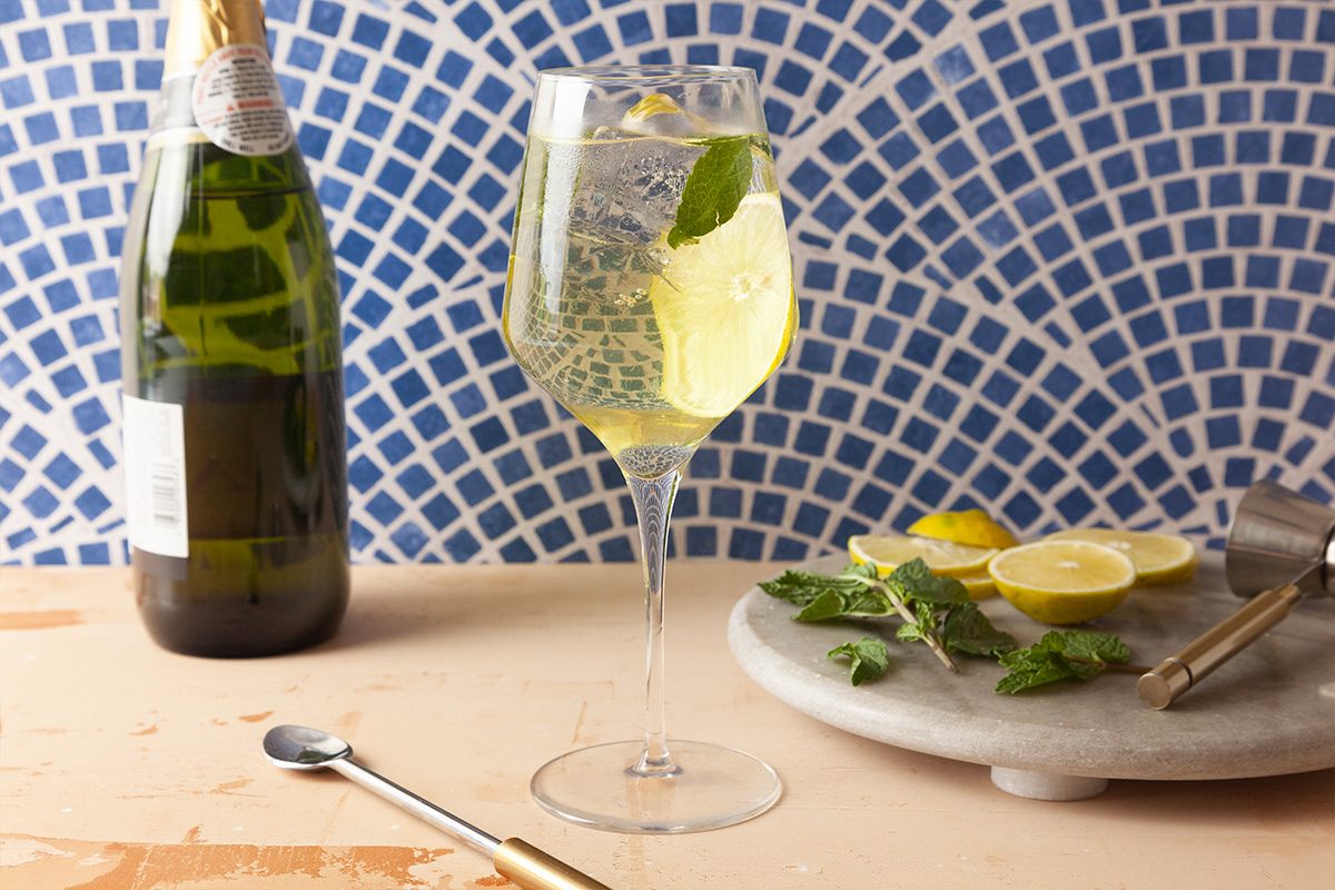 A glass of sparkling white cocktail with ice and mint leaves sits beside a bottle of champagne and a marble tray with sliced lemon and mint, against a blue tiled background.