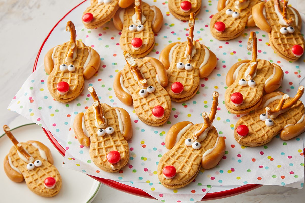 A plate of reindeer-shaped cookies made with peanut butter sandwich cookies, candy eyes, red candy noses, and pretzel antlers, arranged on festive polka dot parchment paper.