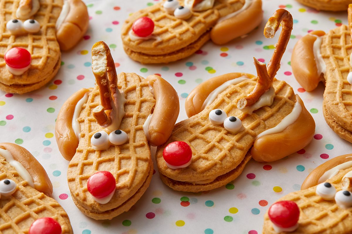 Nutter Butter cookies decorated as reindeer with pretzel antlers, candy eyes, and red candy noses, arranged on a colorful polka dot surface.