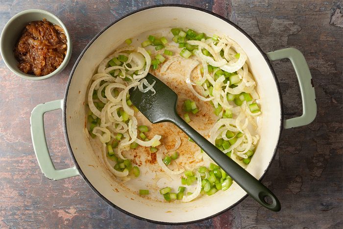 A pot with sautéed sliced onions and chopped green bell peppers sits on a stovetop. A spatula rests inside, and a small bowl containing brown seasoning paste is next to the pot.