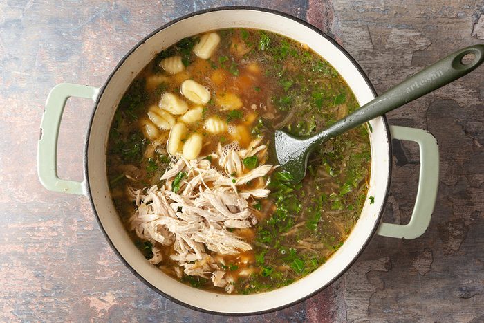 A large pot filled with chicken gnocchi soup, featuring shredded chicken, gnocchi, broth, herbs, and a metal ladle resting inside.