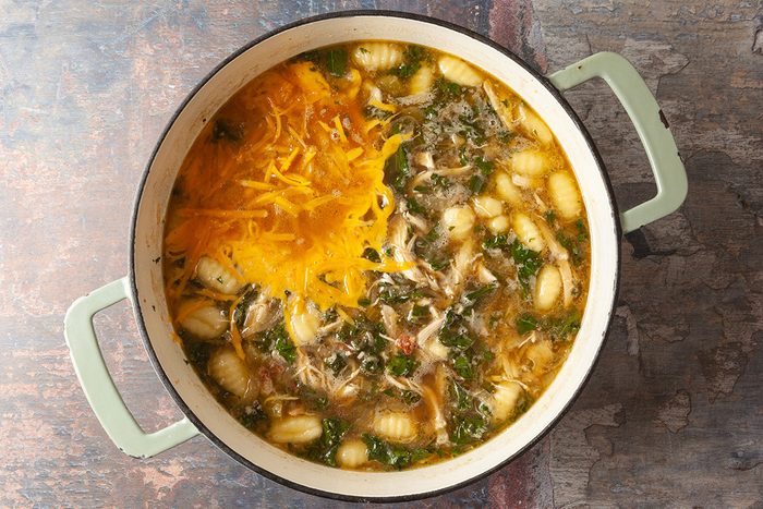 A pot of soup with gnocchi, shredded cheese, leafy greens, and herbs in a light broth, sitting on a rustic surface.