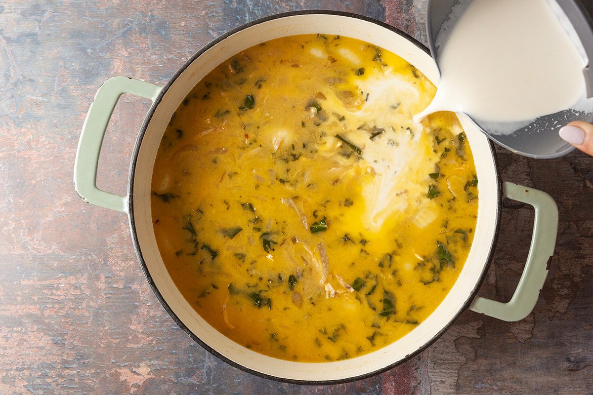 A hand pours a creamy liquid from a measuring cup into a pot of yellow soup with herbs on a rustic countertop.