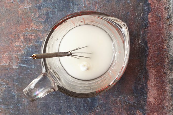 A glass measuring cup filled with a white liquid and a small metal whisk inside, placed on a textured brown and blue surface.