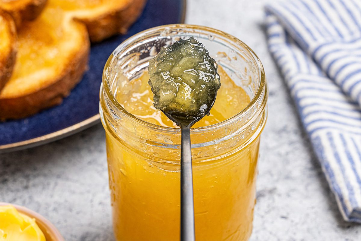 A spoon holding a dollop of yellow marmalade rests above an open glass jar filled with more marmalade. In the background, there are slices of toasted bread and a blue-and-white striped cloth.