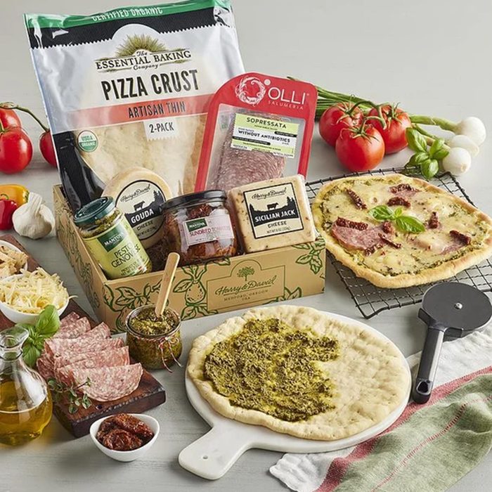 A pizza-making kit with pizza crusts, pesto, salami, cheese, and seasonings, surrounded by fresh tomatoes, garlic, basil, olive oil, and a partially made pizza on a white surface.