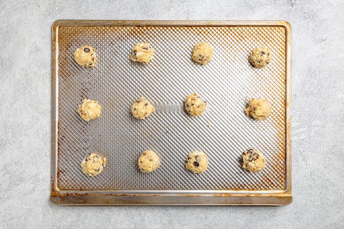 A baking sheet with twelve evenly spaced scoops of chocolate chip cookie dough, ready to be baked, on a light gray surface.