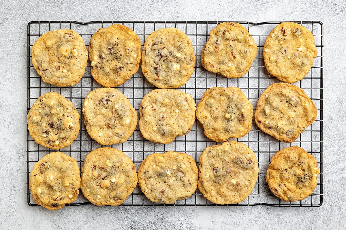 Sixteen homemade cookies with visible chocolate chips and nuts are cooling on a black wire rack placed on a light gray textured surface.