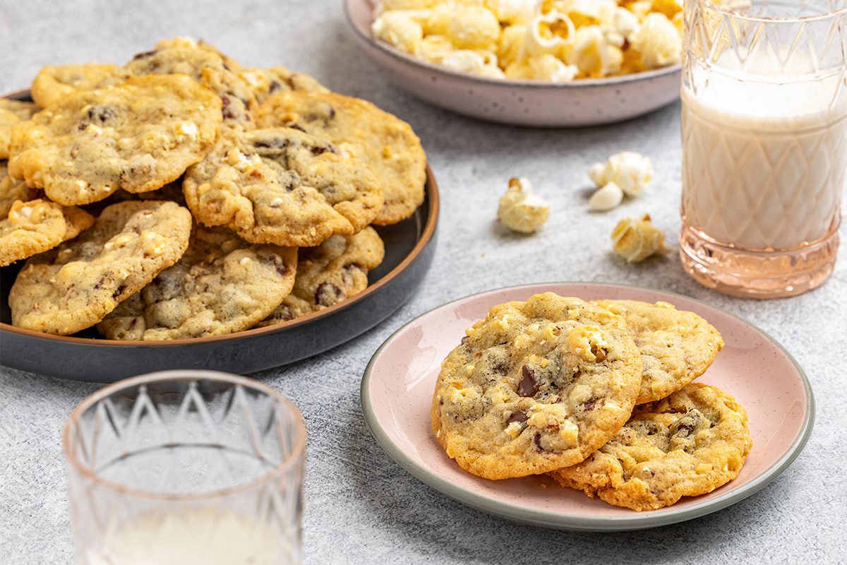 A plate of chocolate chip cookies sits next to a glass of milk, with a smaller plate holding two cookies. In the background, there is a bowl of popcorn and another glass of milk on a light surface.