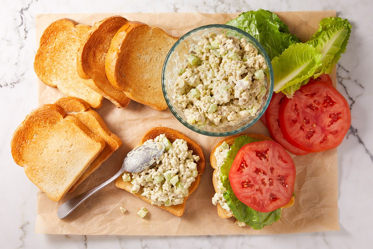 Slices of toasted bread surround a bowl of chicken salad. One slice is topped with lettuce, tomato slices, and chicken salad. A spoon with salad sits on a slice nearby. The scene is set on parchment paper over a marble surface.
