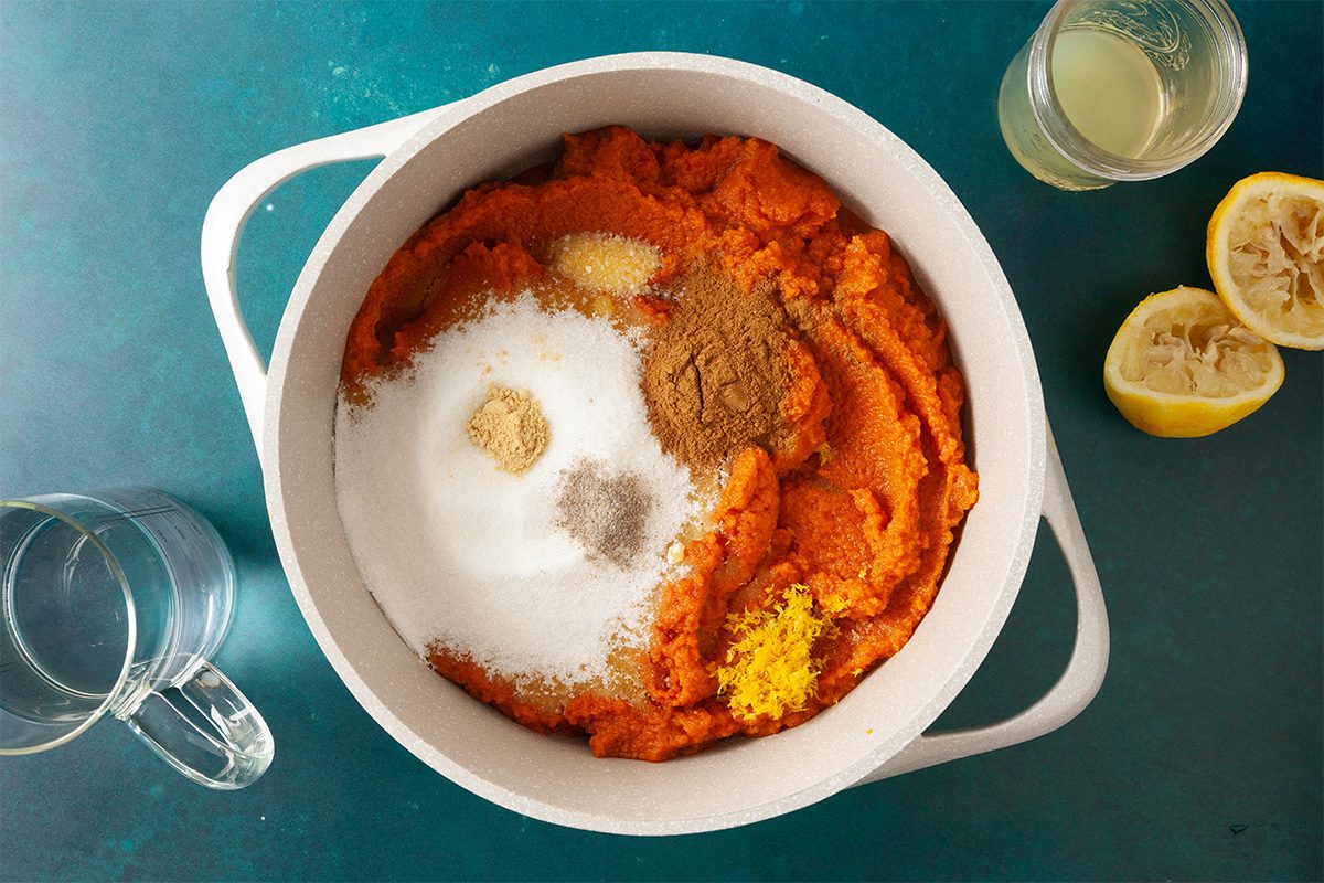 A white pot filled with pumpkin puree, sugar, spices, and lemon zest sits on a teal surface. Nearby are a glass of water, a glass of juice, and two squeezed lemon halves.