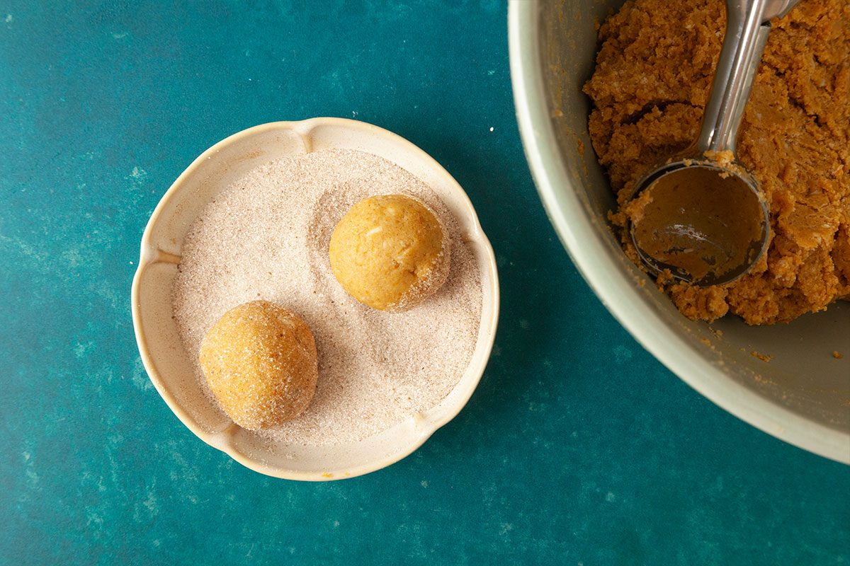 Two dough balls are being coated in sugar on a small plate, next to a mixing bowl filled with more dough and a cookie scoop, all on a teal surface.