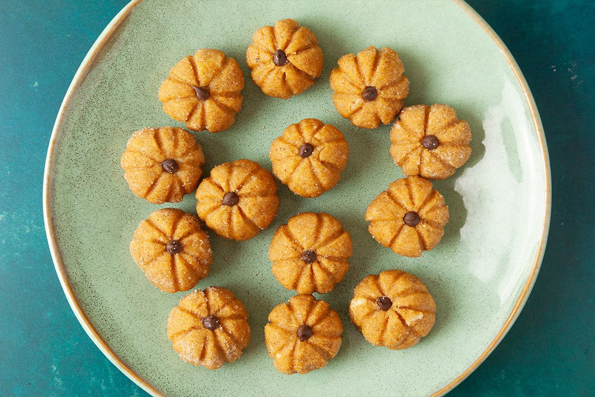 A green plate holds fifteen small cookies shaped like pumpkins, each with ridged sides and a chocolate chip in the center, arranged in a loose pattern on a teal surface.