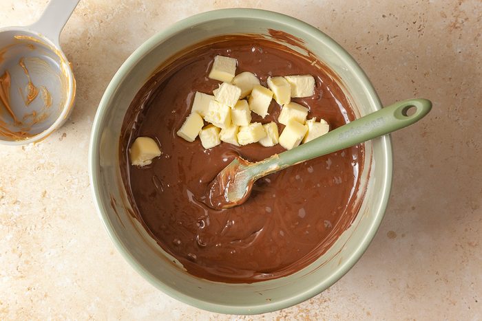 Overhead shot of a green bowl filled with chocolate batter, topped with small cubes of butter; A green spatula rests inside the bowl; while a measuring cup with batter remnants sits nearby on a beige countertop