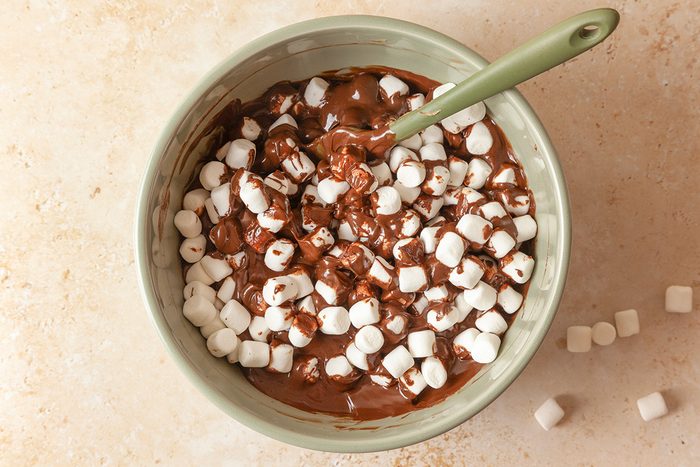 Overhead shot of a green bowl filled with melted chocolate and mini marshmallows, with a green spoon inside; A few marshmallows are scattered across the beige surface beside the bowl