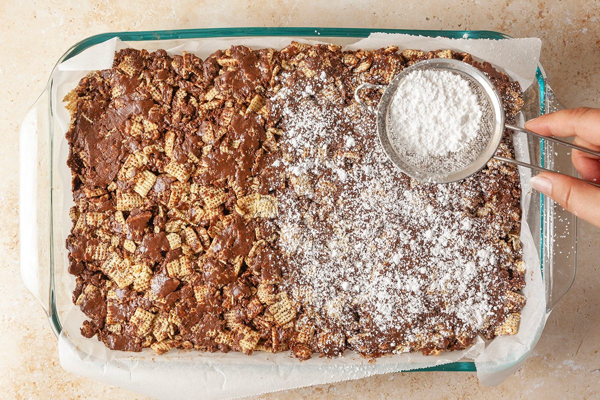 Overhead shot of a hand sprinkling powdered sugar over a chocolate cereal bar mixture in a glass baking dish; with half of the dessert covered in sugar while the other half remains plain