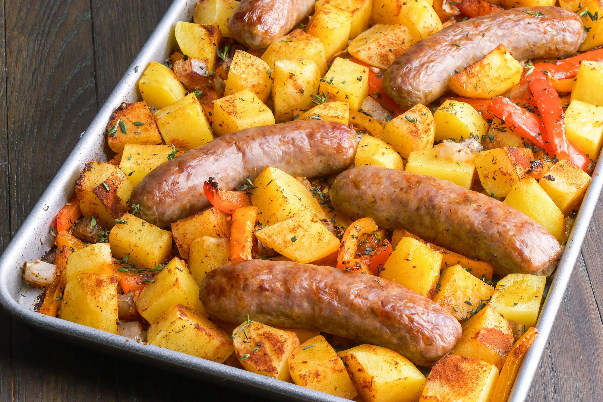 3/4th shot of a sheet pan filled with roasted sausages, diced potatoes, and sliced red bell peppers, seasoned with herbs, on a dark wooden surface