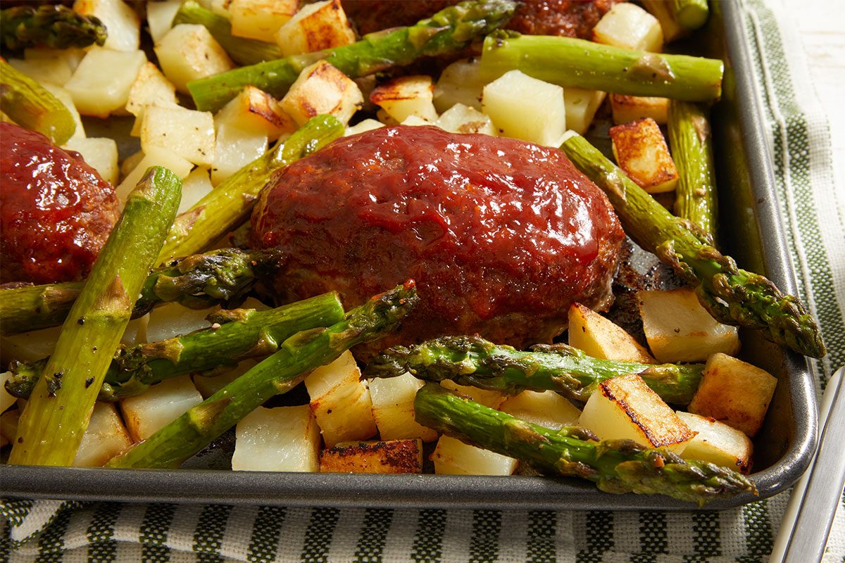 A baking sheet with glazed mini meatloaf, roasted asparagus spears, and diced potatoes, all cooked to a golden brown. The tray is placed on a green and white striped cloth.