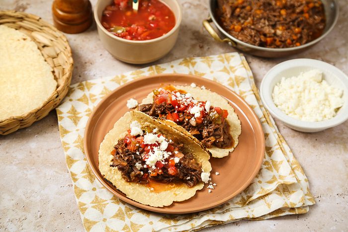 Two beef tacos topped with salsa and crumbled cheese on a tan plate, placed on a patterned napkin. Nearby are a bowl of salsa, a bowl of crumbled cheese, a woven basket with tortillas, and a skillet of beef filling.