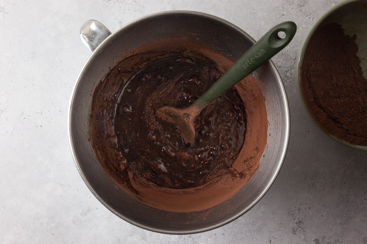 A metal mixing bowl filled with chocolate batter and a green spatula, viewed from above on a light gray surface. Another bowl with cocoa powder is partially visible to the right.