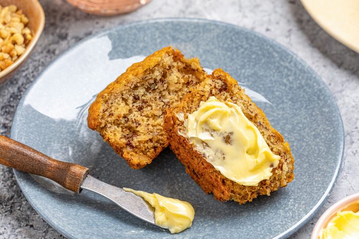 Close-up shot of a halved vegan banana muffin on a blue plate; one half spread with butter and a butter knife resting beside it; placed on a gray surface