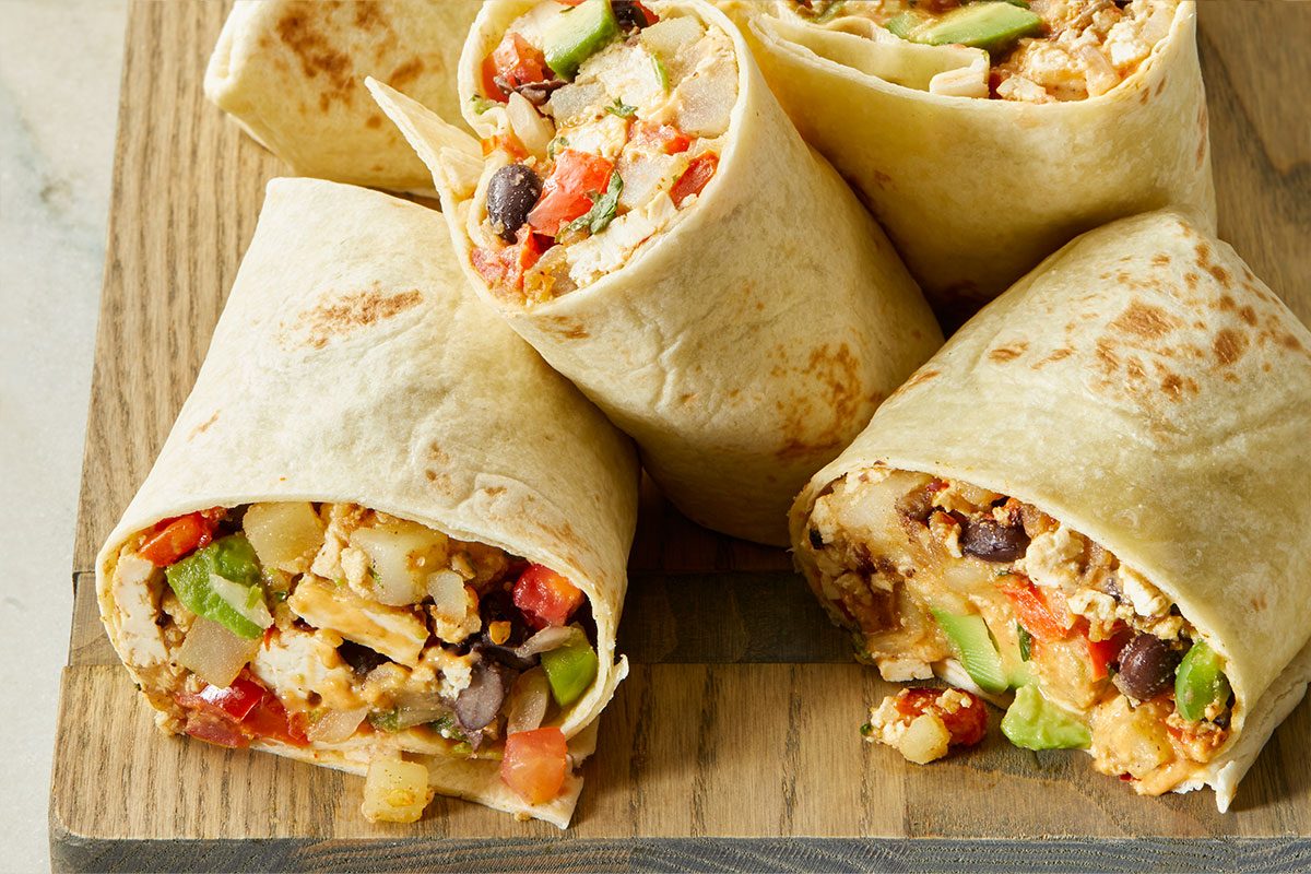 Four burrito halves filled with scrambled eggs, black beans, diced vegetables, and lettuce are arranged on a wooden board, showing their colorful, fresh ingredients inside soft flour tortillas.