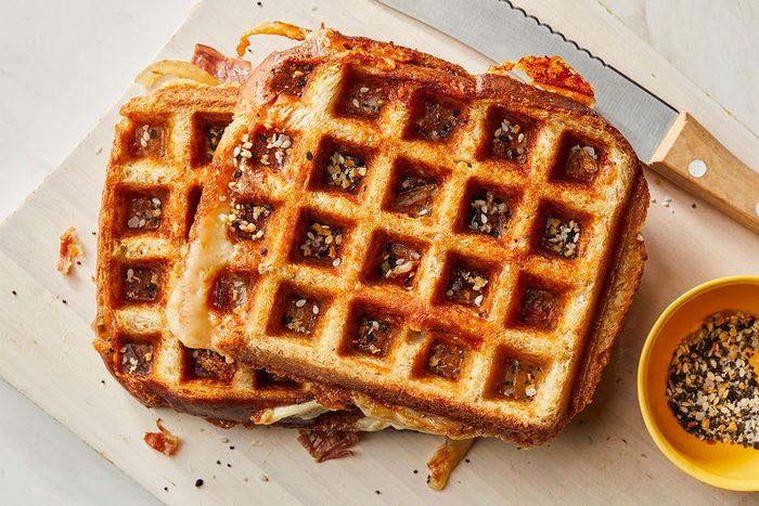overhead shot of Two golden brown waffle sandwiches with melted cheese and seasoning on top rest on a wooden cutting board next to a yellow bowl of seasoning and a bread knife