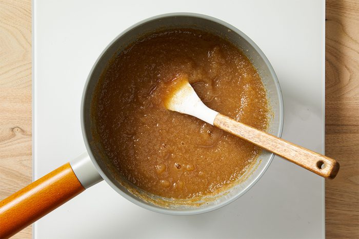 A saucepan filled with thick, brown applesauce sits on a white stovetop. A wooden spoon with a white tip rests in the mixture, which appears smooth with some small chunks. The background is a light wood surface.