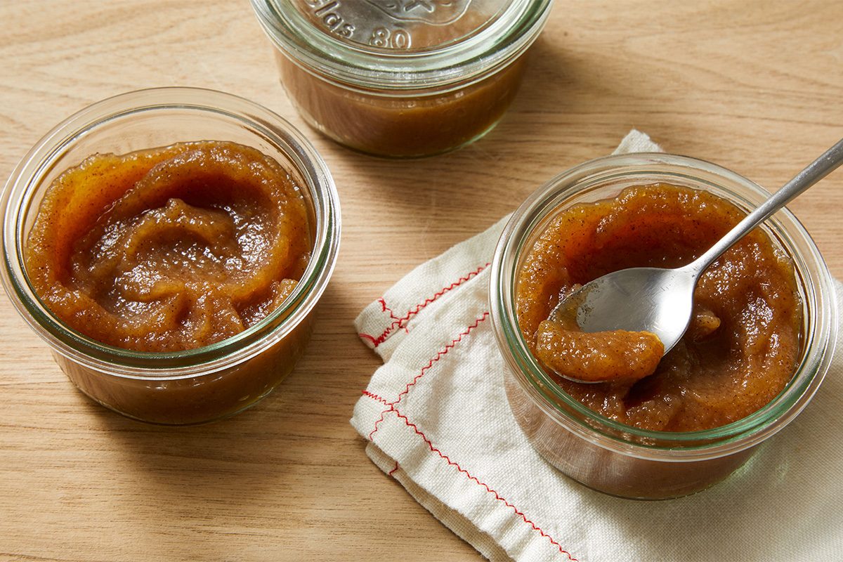 Three glass jars of thick, brown apple butter sit on a wooden surface. One jar has a spoon in it, resting on a folded white cloth with red stitching.