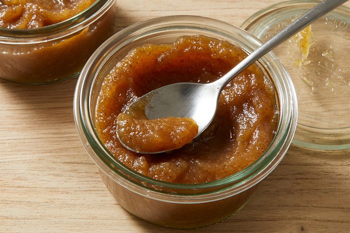 A glass jar filled with thick, brown apple butter sits on a wooden surface. A spoon rests inside the jar, scooping up some of the smooth, glossy spread. Another jar with a lid is partially visible in the background.