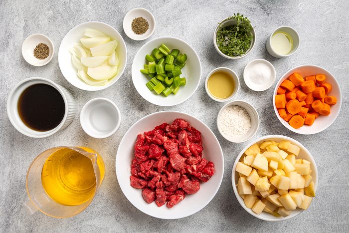 Ingredients for a stew arranged in bowls on a countertop, including chopped beef, potatoes, carrots, celery, onions, broth, seasonings, oil, flour, and herbs.