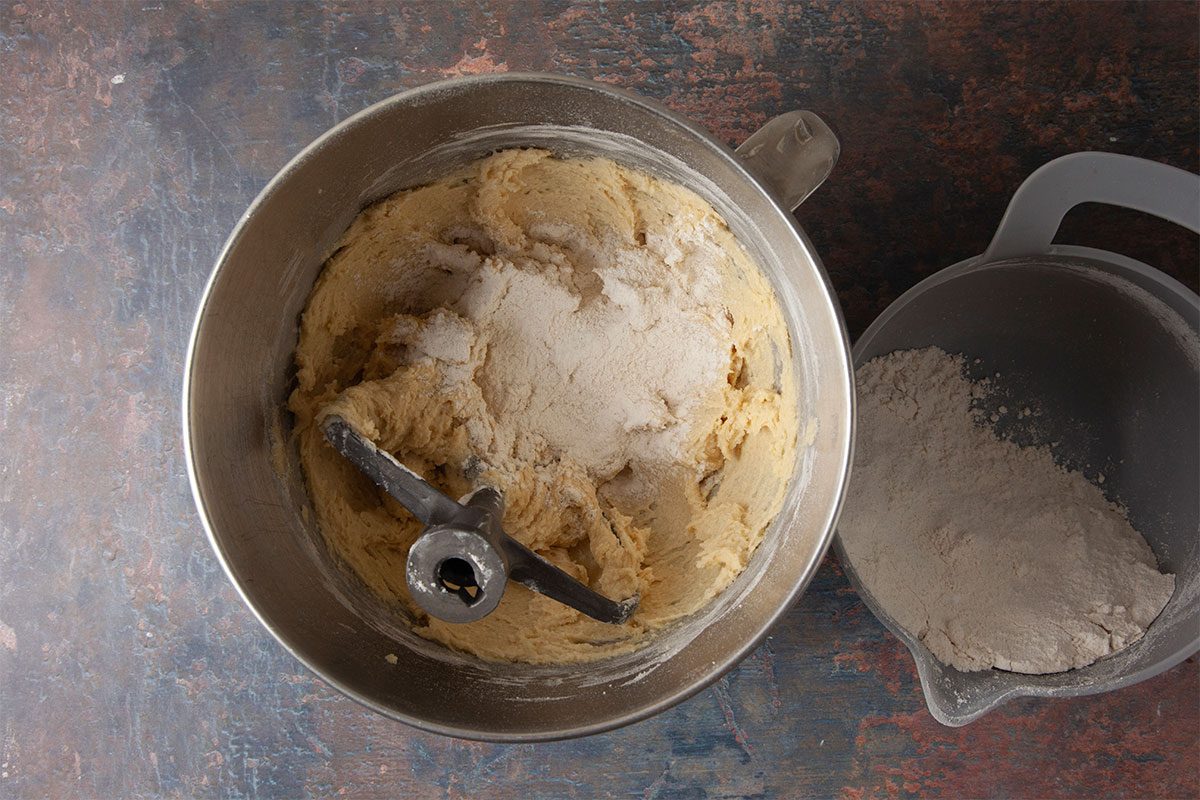 Overhead shot of a mixing bowl filled with creamy batter as flour is being added, placed beside a plastic container of flour on a dark countertop