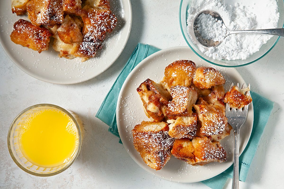A plate of powdered sugar-dusted baked French toast bites, a fork, a bowl of powdered sugar, a second plate of toast, and a glass of orange juice on a white surface with a teal napkin.