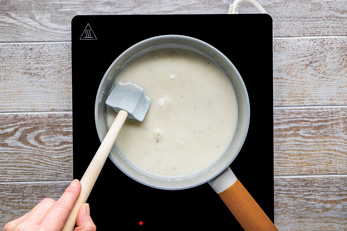 A hand holding a spatula stirs a pot of white sauce on an electric stovetop with a wooden surface underneath.