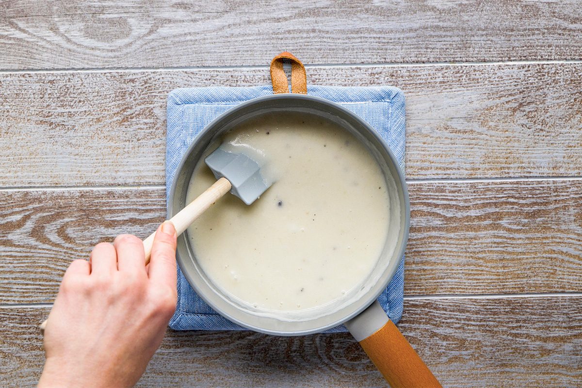 A hand stirs a creamy white sauce with a spatula in a saucepan placed on a blue quilted pot holder, on a wooden surface.