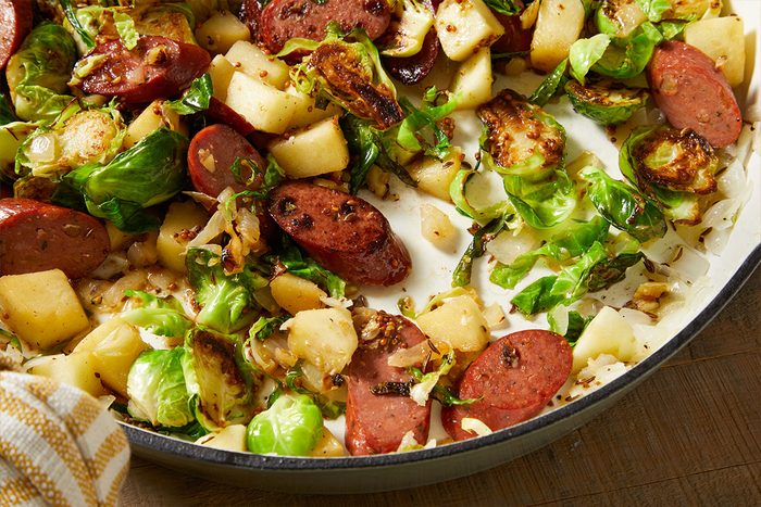 A skillet filled with sliced sausage, Brussels sprouts, and diced potatoes, all cooked and browned, with a striped towel partially visible beside the pan.