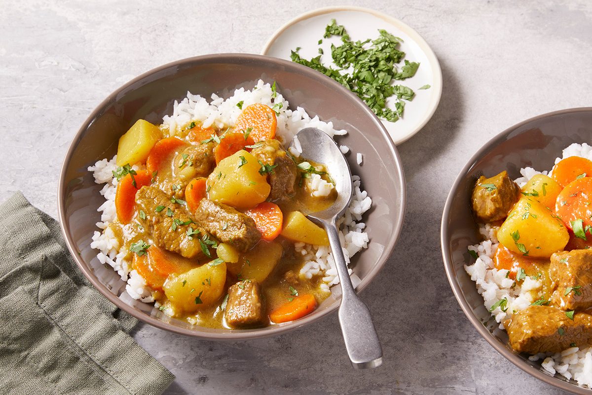 Two bowls of white rice topped with a stew containing chunks of meat, potatoes, and carrots. A spoon rests in one bowl. A small plate of chopped green herbs sits nearby on a gray surface.