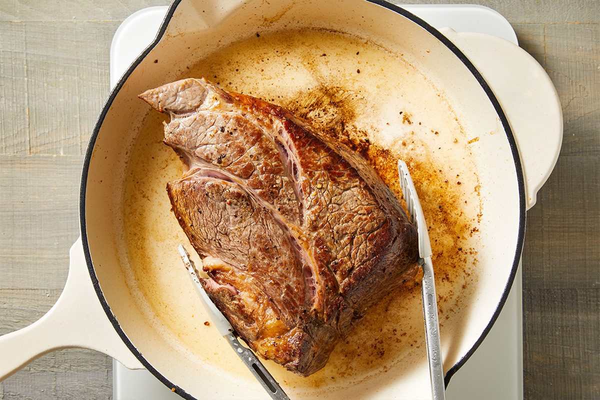 Overhead shot of beef roast searing in a white Dutch oven with tongs resting on the edge.