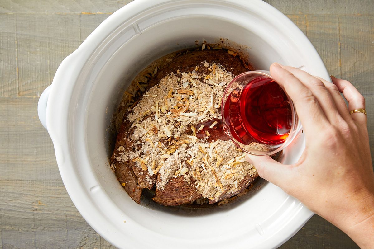 Overhead shot of red wine being poured over seared beef in a slow cooker.